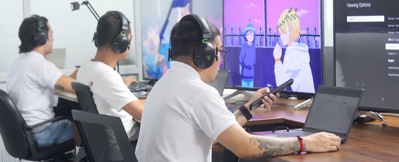 A team of professionals wearing headphones sits at a workstation, monitoring and reviewing anime content on multiple screens. One person holds a remote control while adjusting viewing options on a display, ensuring accurate audio and subtitle settings.