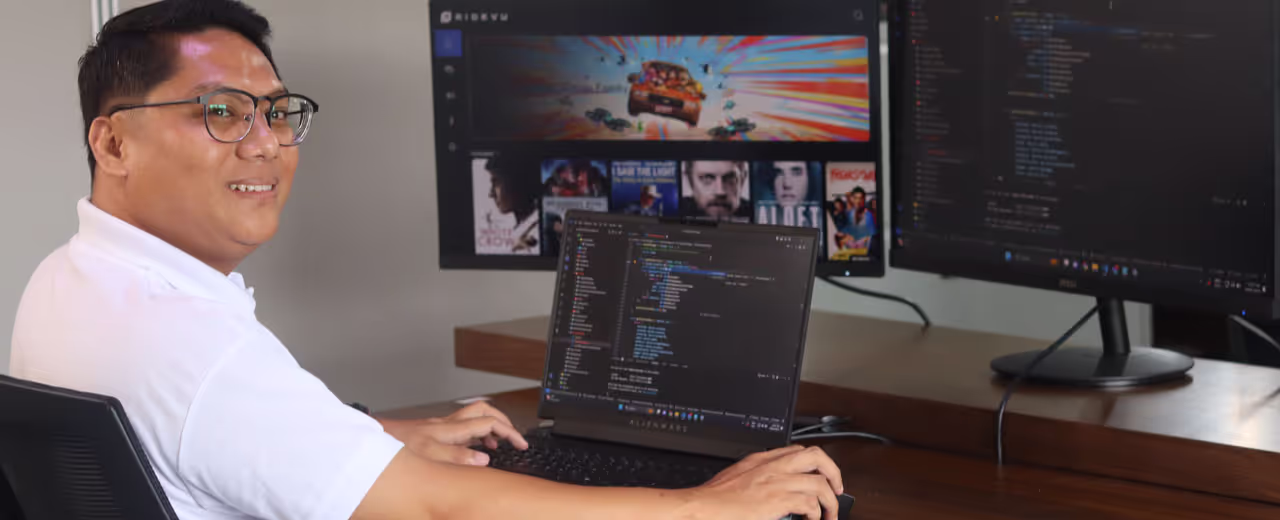 A developer wearing glasses and a white polo shirt works on an Alienware laptop, coding in Visual Studio Code. Two external monitors display additional lines of code and a media streaming interface in the background.
