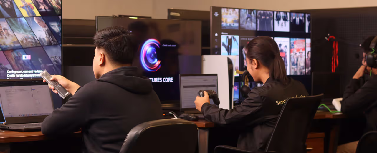 A team of testers works in a dimly lit workspace, evaluating a streaming platform on multiple screens. One tester holds a remote, another uses a game controller, and a third wears a headset while interacting with the system. A PlayStation 5 is visible on the desk, indicating testing across multiple devices.