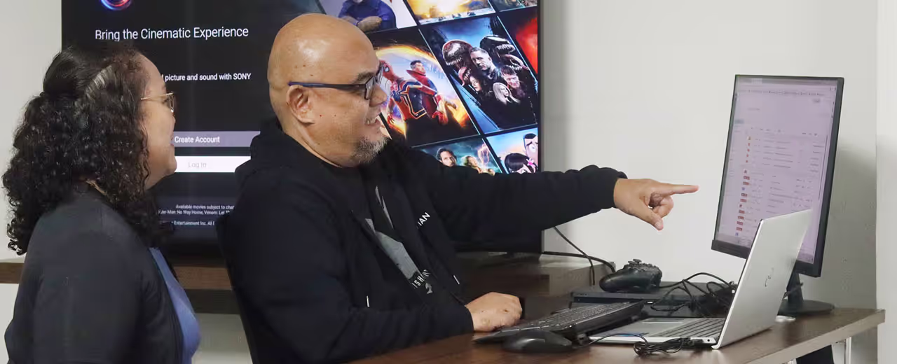 A man wearing glasses and a black hoodie enthusiastically points at a computer screen while discussing something with a colleague. Behind them, a large monitor displays a streaming platform’s interface, showing various movie posters and a 'Create Account' login page.
